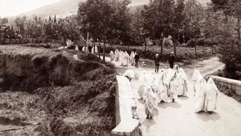 Museum of Archaeology and Anthropology Tlemcen, Algeria. A black and white 19th Century photo showing white-clad women walking back from a cemetery with some crossing a bridge and others walking along a tree-lined road. Behind the first set of women are three men in dark clothing with (possible) Fez hats