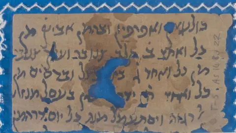 University Library A close-up of a fragment of a medieval manuscript. It has dark writing on a light brown background and is laid on a bright blue background which can be seen through a hole in the manuscript in its middle 