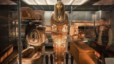 The Fitzwilliam Museum A highly-decorated Egyptian mummy case standing upright in a glass case with decorated sarcophagus to its right and a plain wooden box to its left. A man is looking through the glass at it and other ancient Egyptian artefacts can be seen behind it