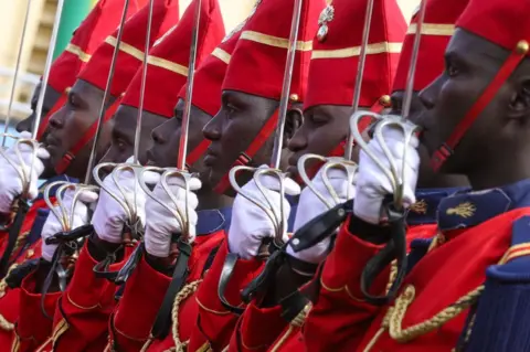 CEM OZDEL / GETTY IMAGES Soldiers dressed in red stand to attention and present their swords at a ceremony in Thiaroye Camp.