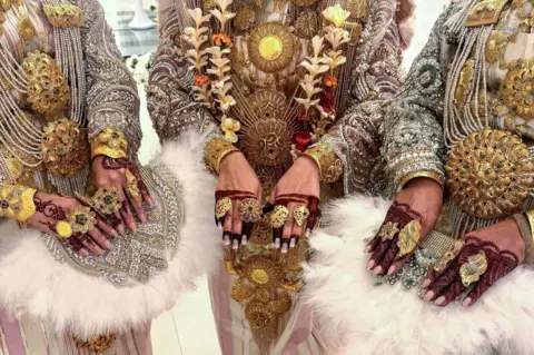 MAHMUD TURKIA / AFP Women wear ornate golden necklaces and rings. On their fingers they have brown-red henna patterns.