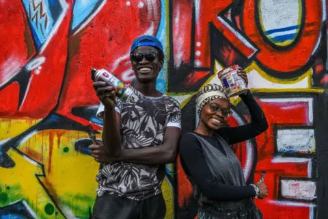 GERALD ANDERSON / GETTY IMAGES A smiling man and woman stand next to a painted wall, carrying their cans of paint.