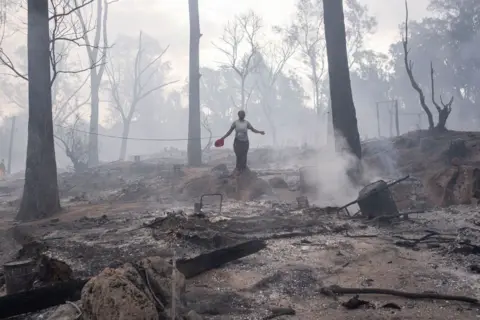 EMMANUEL CROSET / AFP A woman stands among the burnt remains, surrounded by smoke and bare trees.