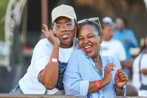 Sharon Seretlo / Getty Images Two women smile and throw peace signs. One sticks her tongue out as she poses for the camera. Behind them is an outdoor stage in Kroonpark Resort - Saturday 7 December 2024