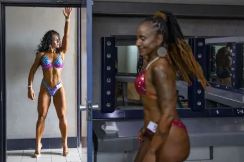 Roberta Ciuccio / AFP Two muscular women in bikinis - one in high heels stretches up on her arms arms backstage at the Kempton Park Civic Centre in Ekurhuleni - Saturday 7 December 2024