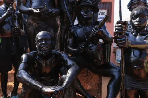 Daniel Beloumou Olomo / AFP Topless men painted in black body paint crouch together. Their expressions are stern and some carry ceremonial weapons in Foumban, Cameroon - Sunday 8 December 2024