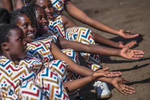 Gerald Anderson / Getty Images Girls in matching multicoloured dressed sit in a row on the ground with with arms outstretched and smiling - Nairobi, Kenya, Saturday 7 December 2024