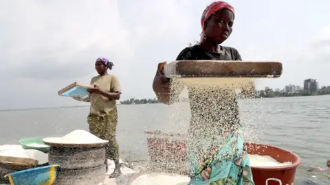 Legnan Koula / EPA Women stand by the sea and sieve cassava grains into large pots. Abidjan's skyline can be seen in the background - Thursday 11 December 2024