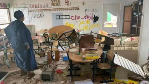 Getty Images A damaged classroom in Mayotte shows tables overturned and children's art on the floor. A woman looks at the scene - with a 