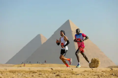 Mohamed Elshahed / Getty Images Two runners run side-by-side during the Marakez Pyramids Half Marathon in Giza, the largest city in Egypt - Saturday 14 December 2024