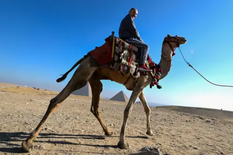 Sayed Hassan / Getty Images A tourist on a camel in Giza, Egypt with the backdrop of the pyramids.