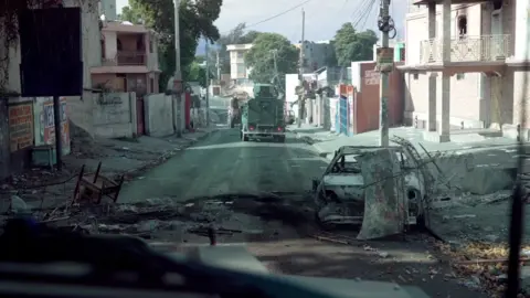 Jack Garland, BBC Burnt out vehicles litter a street in Port-au-Prince, an armoured vehicle can be seen driving ahead. 