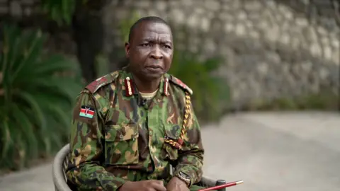 Jack Garland, BBC Godfrey Otunge, in military uniform, sits in a chair. The Kenyan is visible on the sleeve of his uniform shirt. 