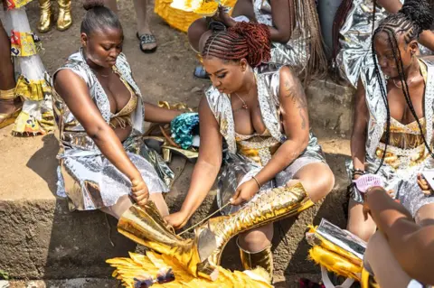 Olympia De Maismont / AFP A woman in silver carnival skirt and waistcoat outfit helps a fellow participant in matching clothes lace up her gold boots - Calabar, Nigeria