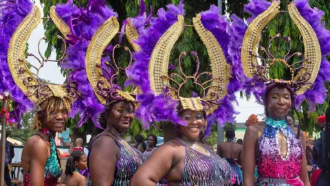 Emmanuel Adegboye / EPA Four women in sequin outfits and purple feathered headdresses attached to manilla like horns at the Calabar carnival smile at the camera