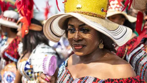 Olympia De Maismont / AFP A woman in a large straw hat and off-the-shoulder outfit holds a pink electric fan as she takes part in the carnival in Calabar, Nigeria