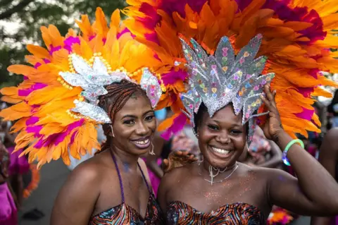 Olympia De Maismont / AFP Two women in headdresses made of silver material and orange and pink feathers smile at the camera at the carnival in Calabar, Nigeria