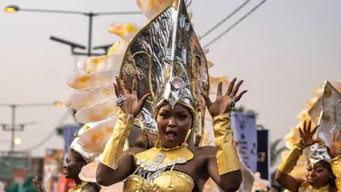 Olympia De Maismont / AFP A performer in a gold outfit with gold glove sleeves and large silver headdress does jazz hands during the Calabar Carnival