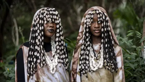 John Wessels / AFP Two women stand near trees. Their clothes are visually striking and their facial expressions are calm. In Dakar, Senegal - Saturday 7 December 2024