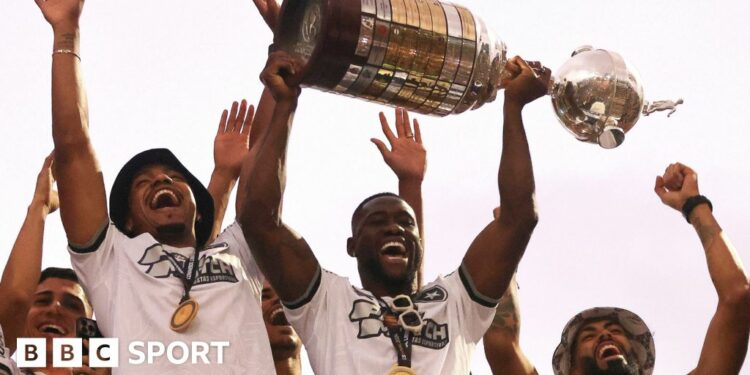 Bastos, wearing a white T-shirt with a gold medal around his neck, lifts the Copa Libertadores trophy above his head while surrounded by four other people lifting their arms in celebration