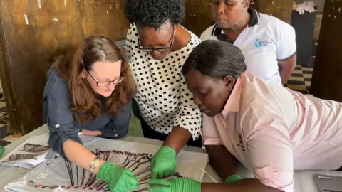 Eva Namusoke Four women are gathered around a table all looking down at glass headband resting on the table on a white cloth. They at Uganda Museum in June 2024. Three of the women, all wearing green plastic gloves, are pointing at or moving the headband. The fourth stands slightly behind them, scrutinising their movements