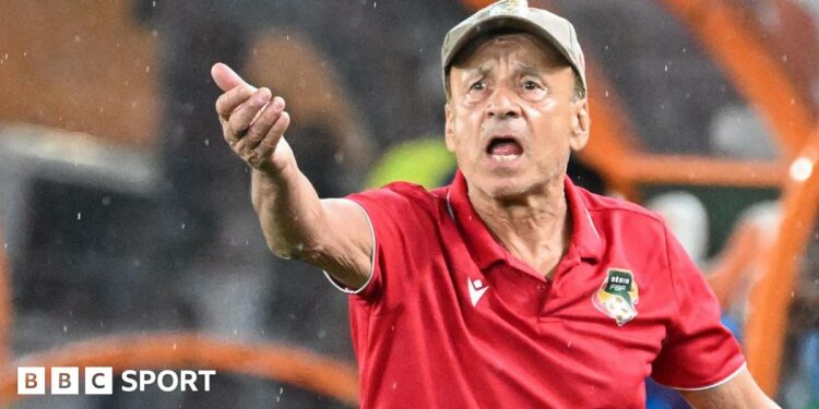 Gernot Rohr wears a beige cap and red polo shirt with Benin football crest on it as he raises his right arm in front of him and gesticulates on the touchline with a look of angst on his face