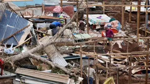 Getty Images Two people survey the damage at a shanty town in Mayotte. A fallen tree and sheets of metal strewn over a hill with damaged wooden frames 