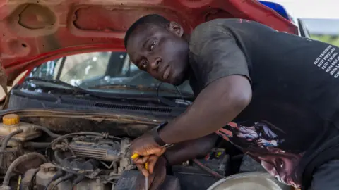 Ernest Ansah / BBC Nathaniel Qainoo, dressed in black, fixes the engine of a car