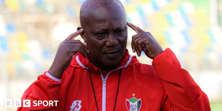 Kwesi Appiah, wearing a red tracksuit top with a Sudan FA logo on the left breast, points his index fingers to his temples