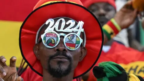 AFP A Ghanaian football fan wearing glasses that say 2024 on them. He has a hat with a red brim.