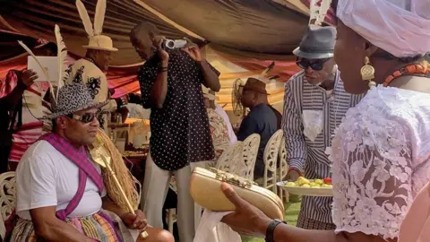 Adaobi Tricia Nwaubani Pascal Okey Adizua is seated, wearing a leopard print hat with two feathers attached to it. He is being approached by a someone carrying  a plate of food and a woman who is attending his Ogbuagu ceremony