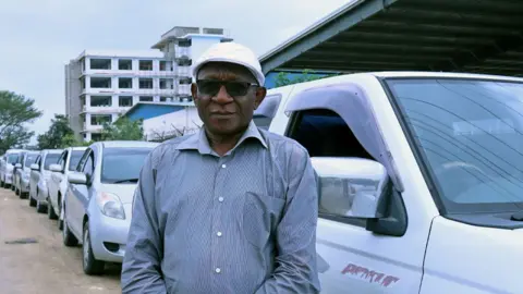 A man in a white cap, dark glasses and shirt stands in front of a line of vehicles.