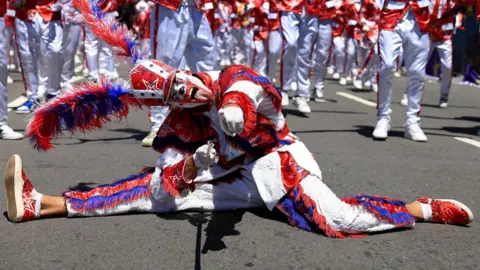 Esa Alexander / Reuters A man wearing a white, red, and blue costume with an elaborate headgear poses in a split position with his tongue sticking out at the Tweede Nuwe Jaar festival - Saturday 4 January 2025