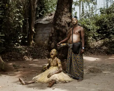 Marco Longari / AFP A man, standing, drizzles gin over a young person sitting on the floor, who is covered in red palm oil mixed with maize flour, giving it a golden hue at a ceremony at the at the annual Voodoo festival in Benin, Ouidah - Thursday 9 January 2025.