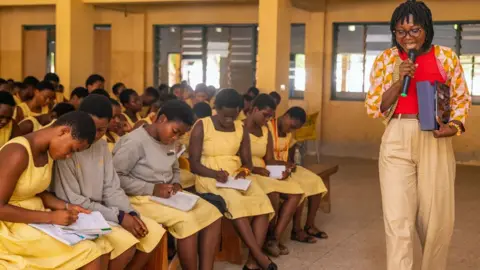 Angela Tabiri Dr Angela Tabiri walks and talks holding a microphone during a class and Innovate Her outreach event at Accra Girls Senior High School. The students, all in yellow dress uniforms, take notes as they sit on benches in a classroom.