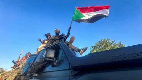 Reuters Men in camouflage,  one of whom is waving a flag, are in a land cruiser in Wad Madani in Sudan on 12 January 2025