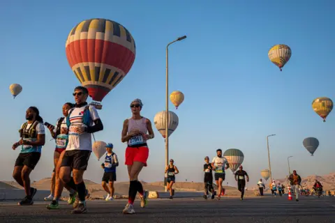 KHALED DESOUKI / AFP People in running gear and with numbers attached to their chests jog on a road. Hot-air balloons rise in the background.