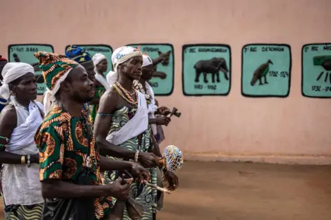 OLYMPIA DE MAISMONT / AFP People wearing matching clothing sing and dance in procession. Behind them is a pink wall which is painted with symbols in turquoise and black.