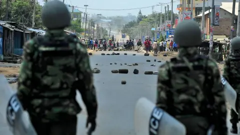 AFP Two Kenyan riot-police officers with their backs to the camera holding shields as people stand near a barricade in Kisumu during protests after the announcement of election results on 9 August 2017