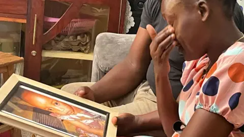 Gladys Kigo / BBC Samantha Pendo's parents sitting on a coach and looking at a framed photo of her taken shorty before her death - Kisumu, Kenya, January 2025