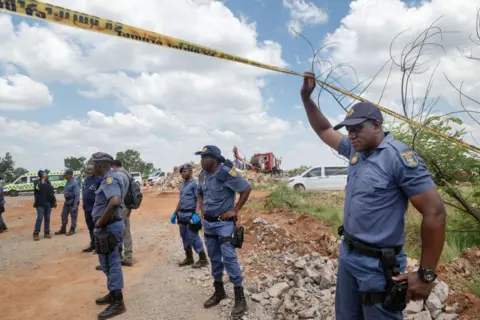 Getty Images Policemen stand on a dirt road as one in the foreground lifts his arm up to hold some police tape aloft.