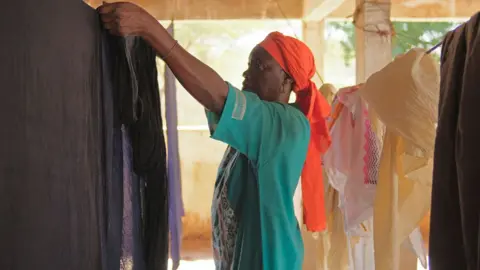 A woman in a green dress and red scarf hangs up dyed black material on a line in Mbacke Kadior