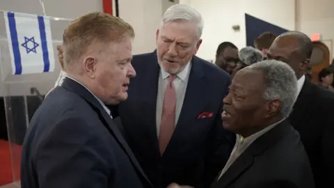 Global Crusade with Kumuyi/Kevin Smart Media William Kumuyi in conversation and shaking hands with Jim Garlow next to a lectern that has the Israeli flag on it.