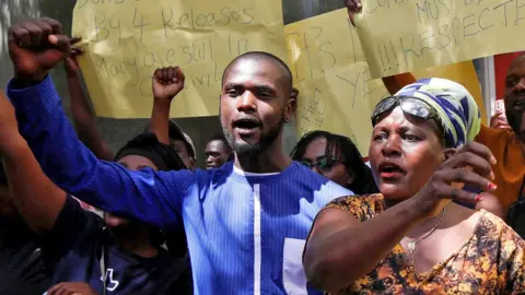 Reuters A of group of Kenyan protesters holding yellow hand-written posters aloft calling for the release of people who have been abducted.  Several - including a young bearded man in a blue collars shirt and a woman wearing a brown patterned V-neck dress, necklace, blue-and-white headscarf and sunglasses on her forehead in close-up - have their fists raised.  