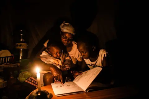 GERALD ANDERSON / GETTY IMAGES A young woman sits with a child and an infant at a table, reading from a school exercise book.