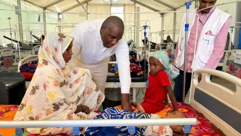 David Lammy in a white shirt bending over a bed wher a mother sits with a child and baby at a MSF clinic in Chad. A MSF medic stands nearby