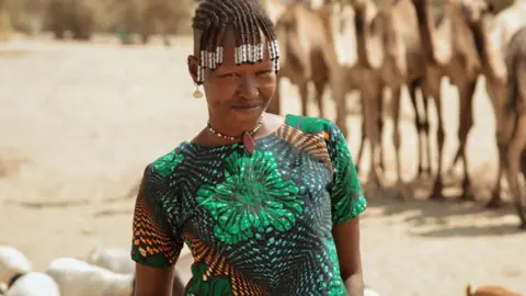 FILMCREW Apuu Mourrine wearing a patterned green top, bead necklace and beads in her braided hair smiles at the camera. Behind her can be seen camels and other livestock