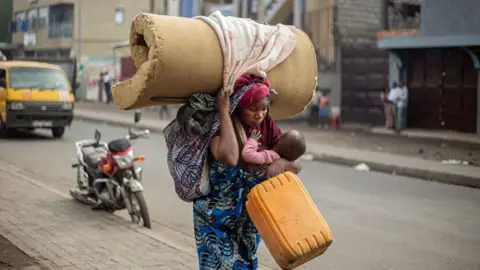 AFP A woman wearing a printed blue wrap walks, slightly hunched forward as she holds a baby, a yellow jerry can and a rolled foam mattress on her head