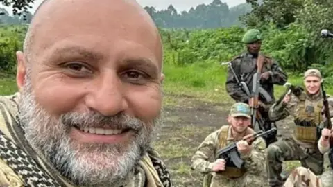 Vasile Badea family Vasile Badea, taking a selfie whilst in DR Congo. He has a grey beard, closely cropped, nearly bald head and a chequered scarf. Behind him to the left can be seen other a few other mercenaries along with a member of the Congolese armed forces - all posing with guns.