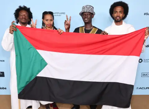 CINDY ORD / GETTY IMAGES Four people stand in a row at a photocall, holding up the Sudanese flag.